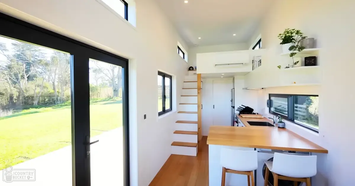 A modern kitchen interior with a wooden bar counter, white stools, shelves holding plants and decor, and sliding glass doors leading to an outdoor garden under bright midday sunlight.