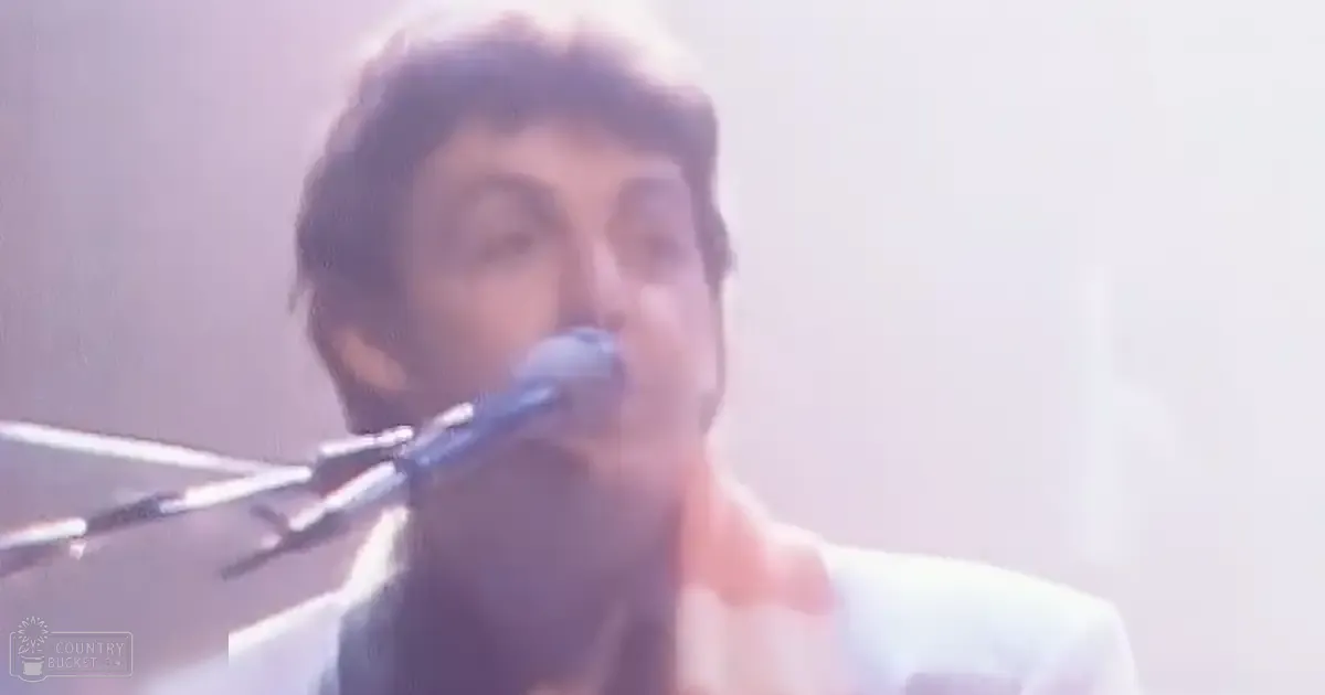 An adult male singer wearing a white tailored suit jacket, singing into a microphone in an indoor concert setting.
