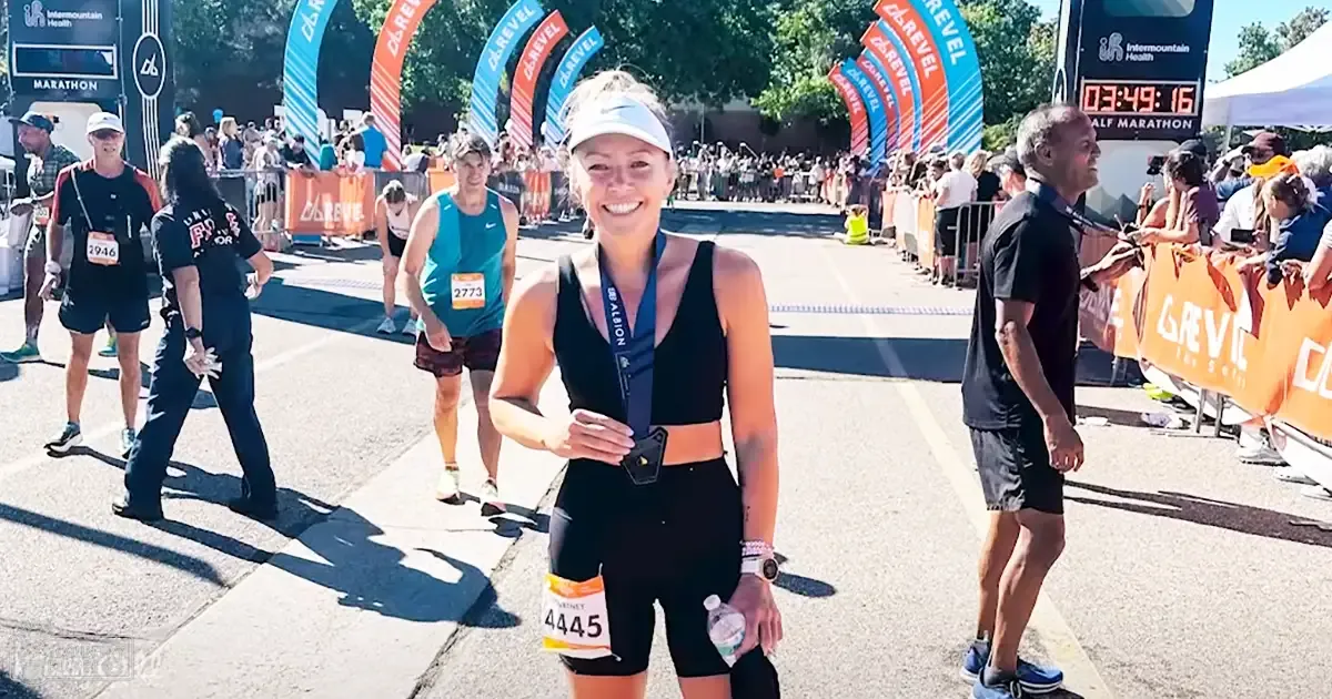 A young adult female finishes a marathon in an outdoor setting with trees and clear skies. She wears a black athletic top with cut-out design and shorts, medal around her neck, wristwatch, bracelet, and holds a water bottle.