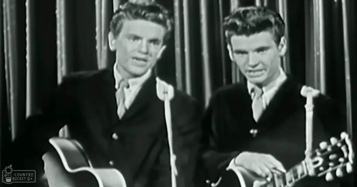 Two young adult males in a studio setting, wearing dark-colored suit jackets with collared shirts; microphones and curtain-like elements suggest a recording or performance environment.