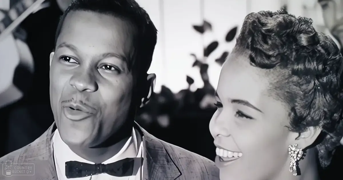 An adult male and female couple dressed formally indoors, with the man wearing a mid-20th century suit and bow tie, and the woman in an upturned collar dress adorned with a decorative accessory.