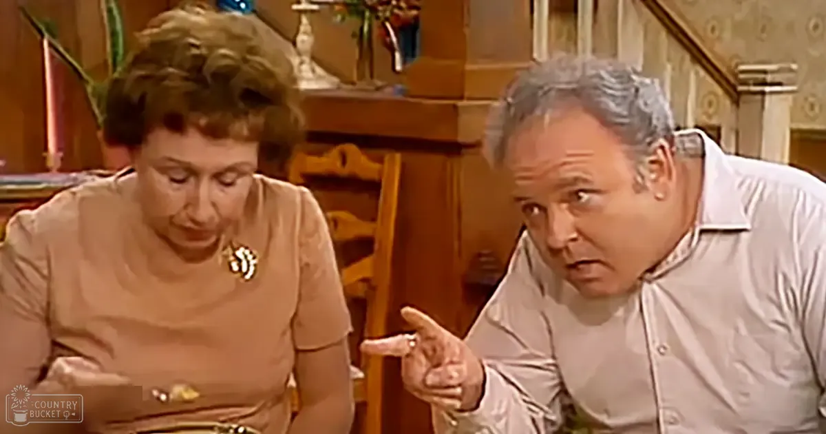 Middle-aged man in light-colored shirt, middle-aged woman wearing floral top with belt and rounded neckline indoors, surrounded by furniture and decorative items.