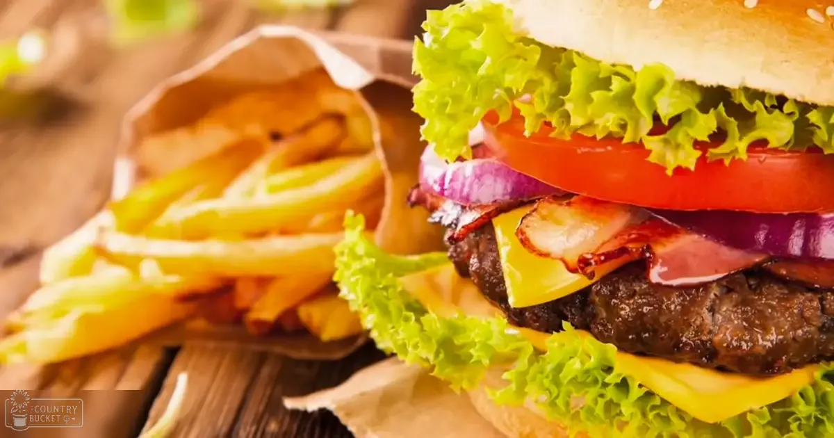 Close-up gourmet burger with lettuce, tomato, bacon, red onions, and cheese served with golden fries on a rustic wooden table in a casual dining setting.