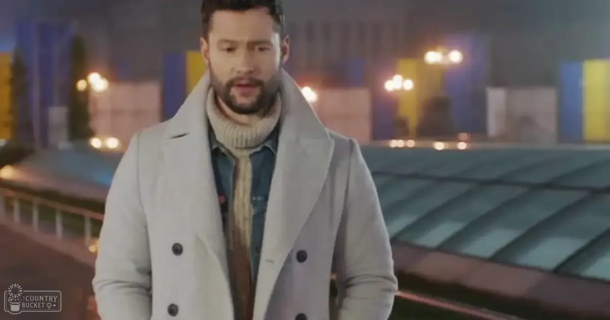 An adult male wearing a light gray double-breasted coat with black buttons, cream or beige high-collared turtleneck sweater, and denim shirt underneath, standing on an urban rooftop at night.