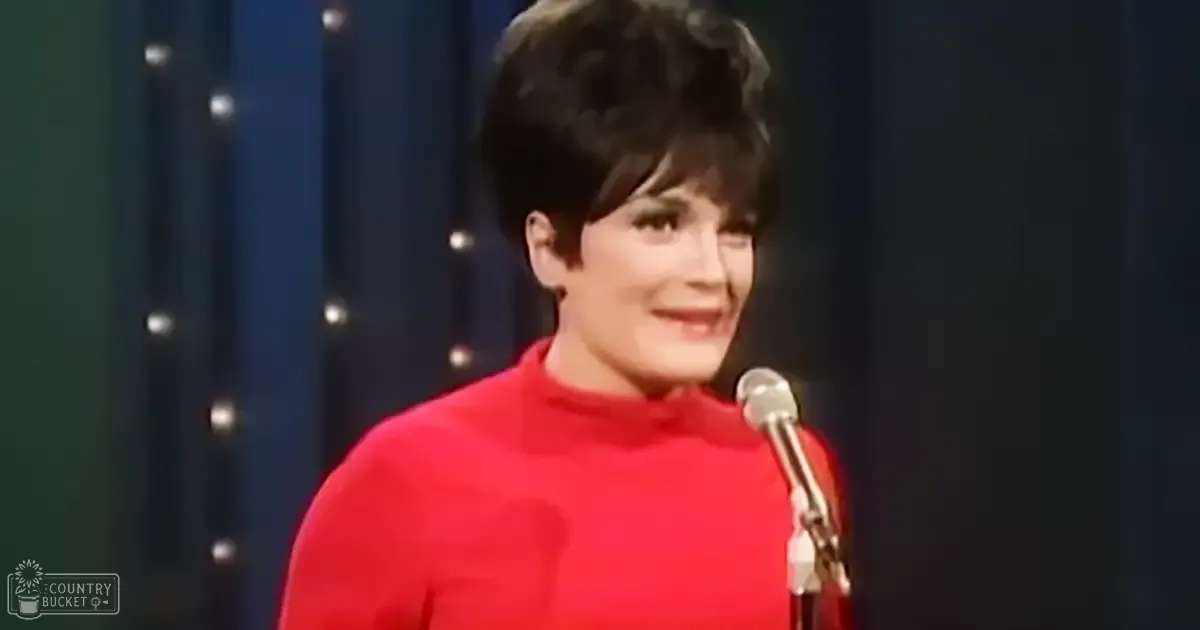A middle-aged adult female performer in a red turtleneck sweater, likely speaking or singing into a microphone at an indoor stage setting.