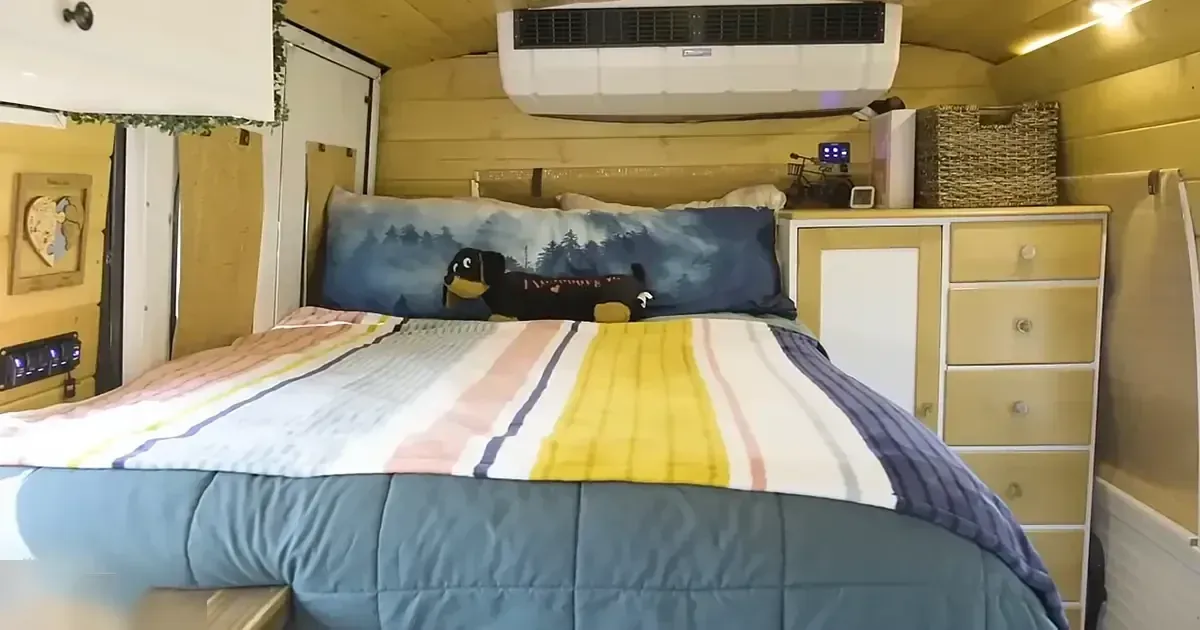 Cozy bedroom setup inside an RV or camper van, with a bed with blue quilt and striped pink, yellow, white blanket, wooden dresser, small shelf, air conditioning unit, storage baskets. Daytime natural light.