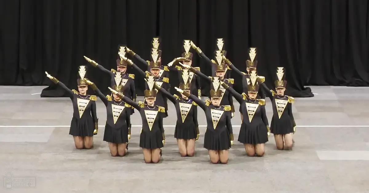 Young adult female dancers in a gymnasium wearing black and gold uniforms with triangular embellishments on the chest and tall hats adorned with golden tassels, kneeling against a black curtain backdrop under artificial lighting.