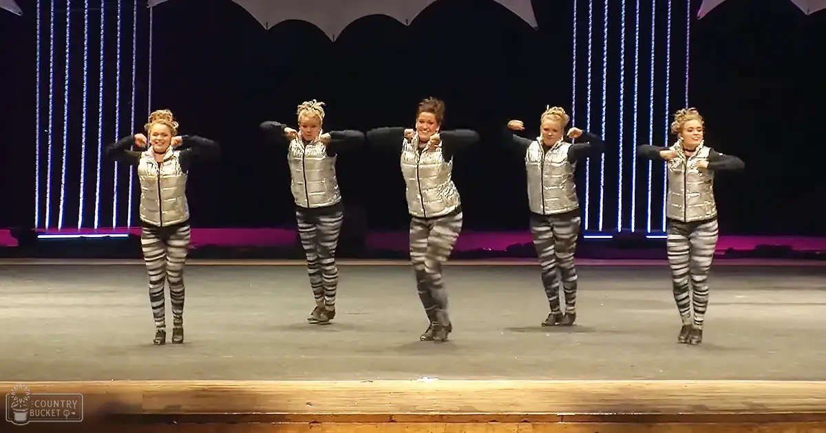 A group of young adult female performers dressed in shiny silver vests over black shirts and patterned leggings stand on a stage with vertical light strips, possibly at an evening school or community event.