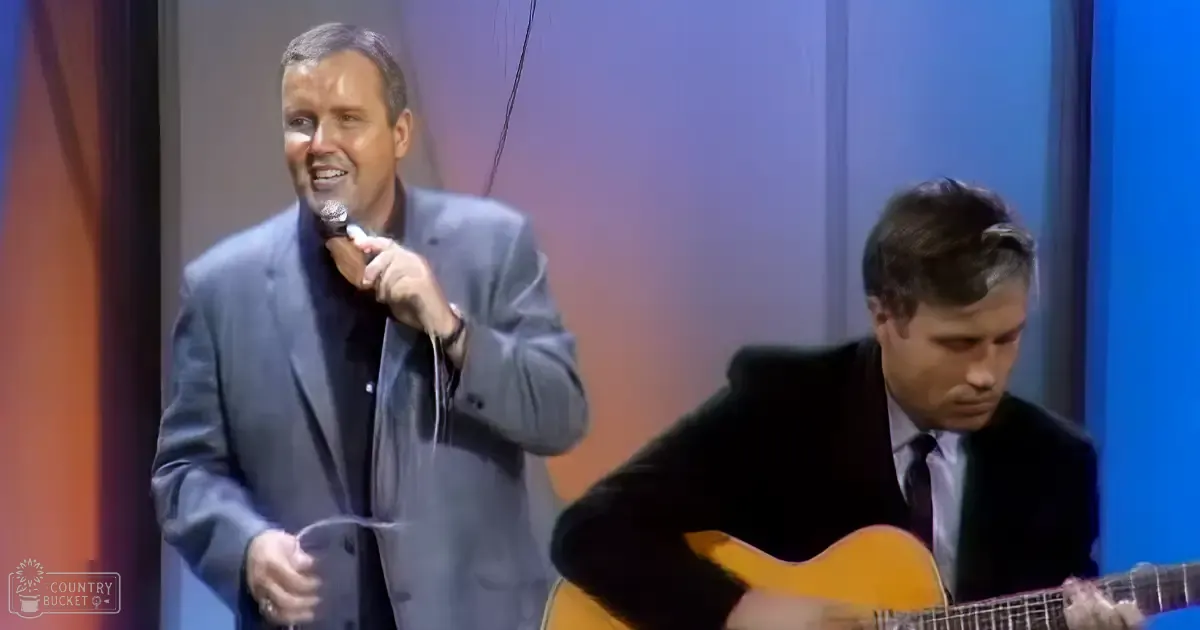 An adult male singer wearing a light gray suit jacket performs into a microphone, accompanied by another middle-aged man playing an acoustic guitar on stage under blue and purple lighting.