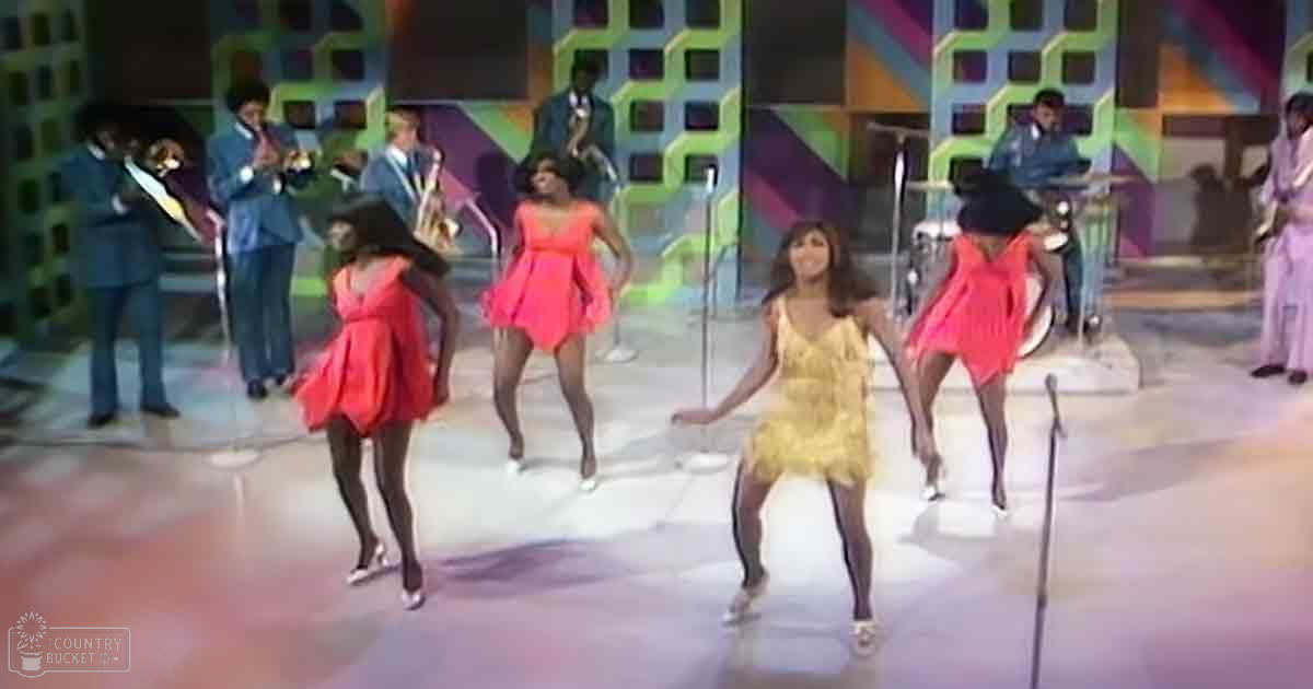 A dance performance with young adult female dancers in vibrant dresses set against a colorful geometric backdrop with musicians playing instruments, suggesting a lively 1960s or early 1970s music show.