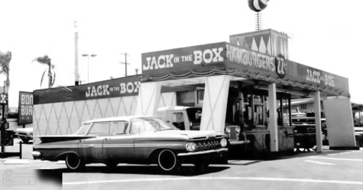 Classic car from mid-20th century parked outside vintage Jack in the Box restaurant with palm trees in background under natural daylight, suggesting summer or spring.