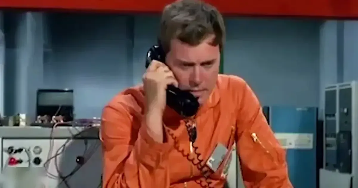 An adult male in an orange jumpsuit with ruffled collar and lanyard, holding a telephone receiver, set against industrial equipment in what appears to be a control room or laboratory.