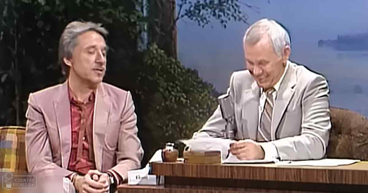 Two individuals in a talk show setting, one wearing a light pink blazer over a red plaid shirt, the other in a gray suit jacket with a white striped shirt and tie.