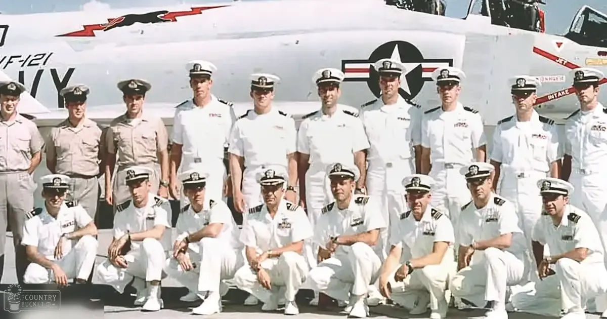 A group of naval personnel in white uniforms with epaulets, seated and standing on an aircraft carrier deck under clear skies.