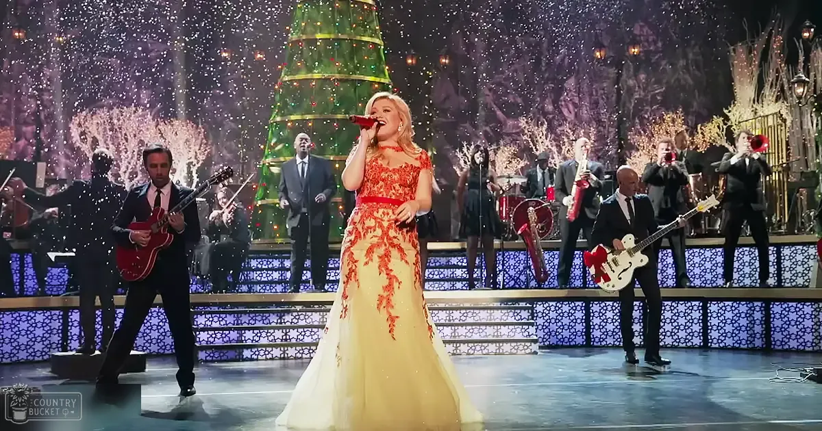 A woman singing in front of a Christmas tree, wearing a long gown with red and yellow patterns, accompanied by musicians in formal attire; indoor holiday performance setting.