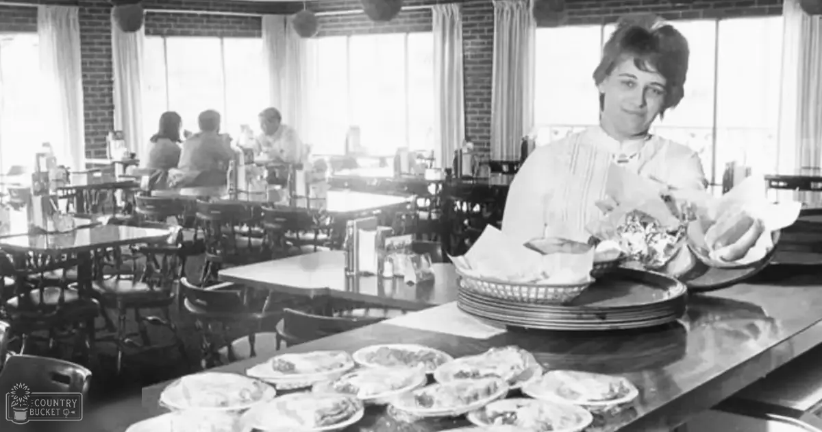 An adult female in mid-20th century attire working at a diner with floral-patterned plates and simple furniture, natural light streaming through large windows on a spring or summer day.