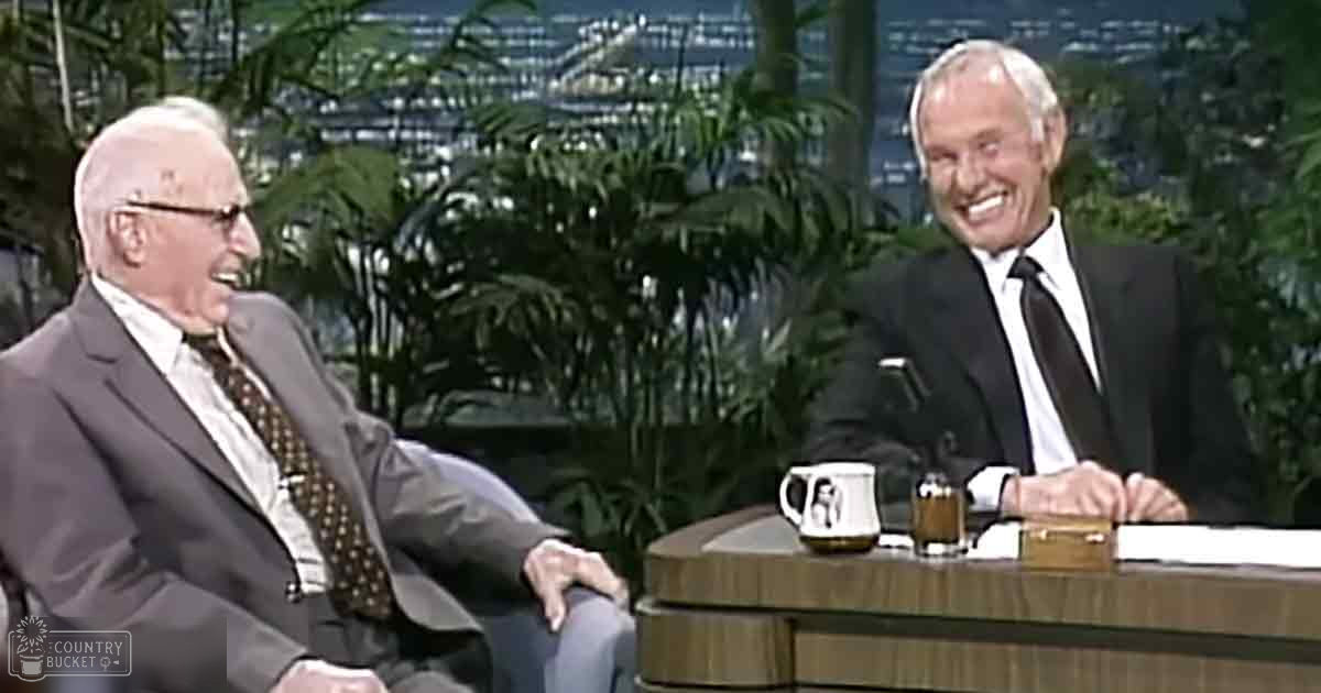 Talk show studio scene with two older adult males seated at a desk, surrounded by indoor plants and large windows showing an out-of-focus urban view.
