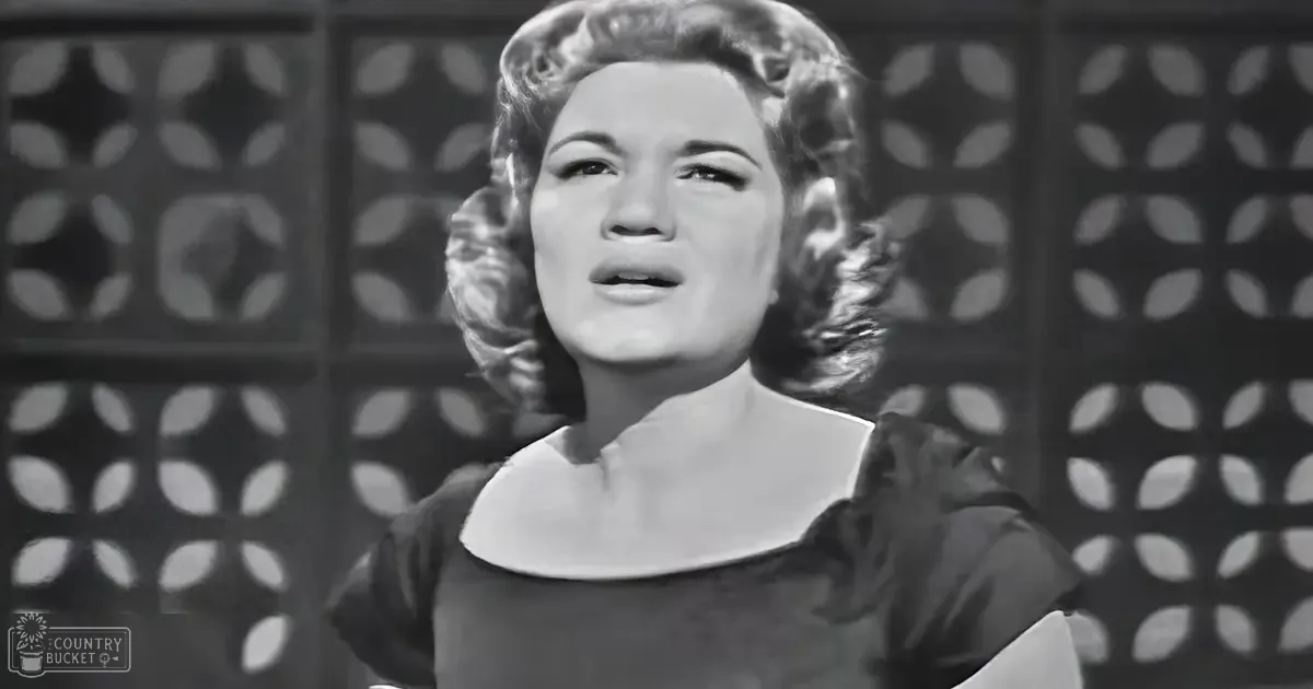 A middle-aged woman in a dark dress with short sleeves and a round neckline stands indoors against a geometric-patterned backdrop under well-lit conditions.