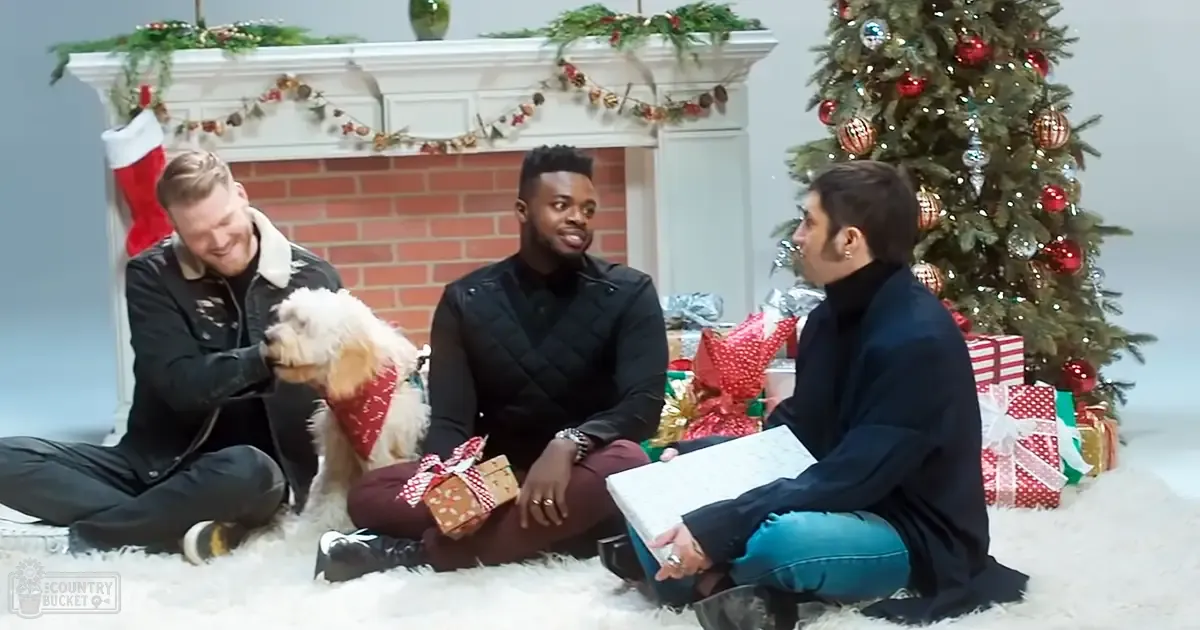 Adult males in festive holiday attire gather around a Christmas tree, holding wrapped gifts and wearing quilted jackets with accessories like watches and rings.