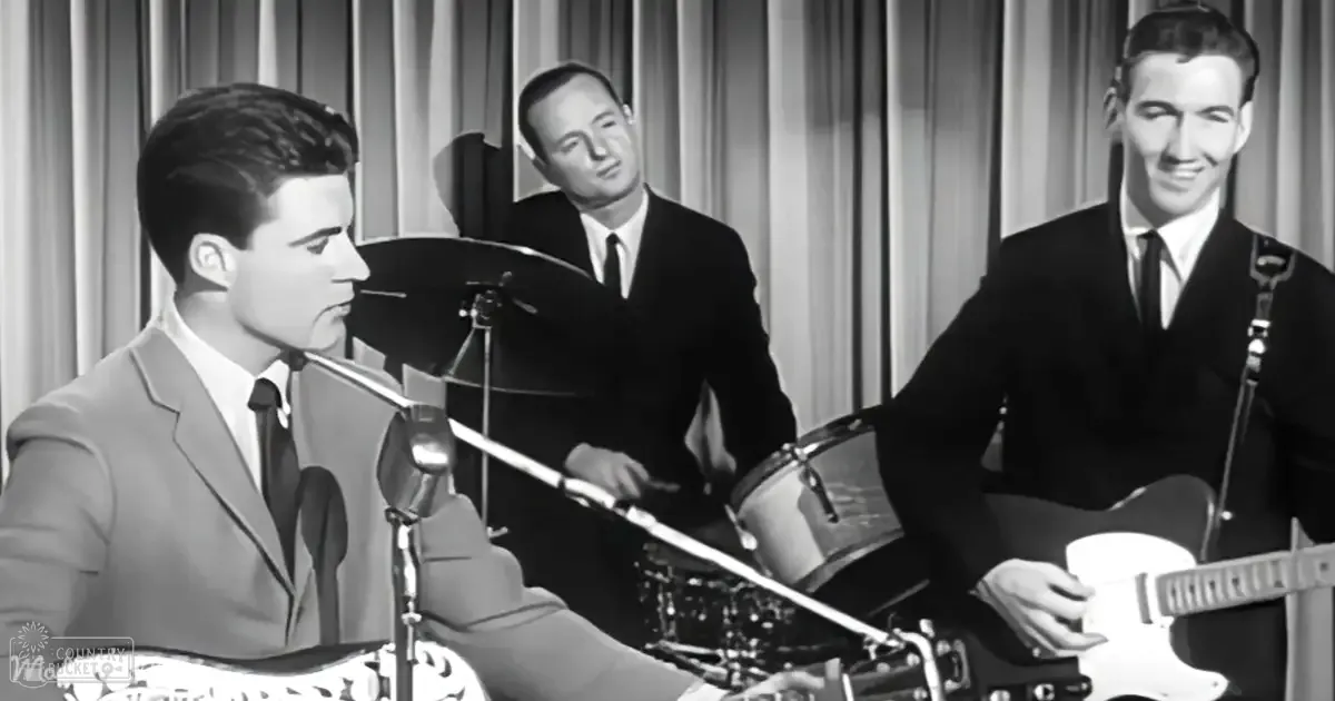 Musical performance with young adult male in light-colored suit jacket and dark tie standing at a drum set in an indoor setting with vertical blinds.