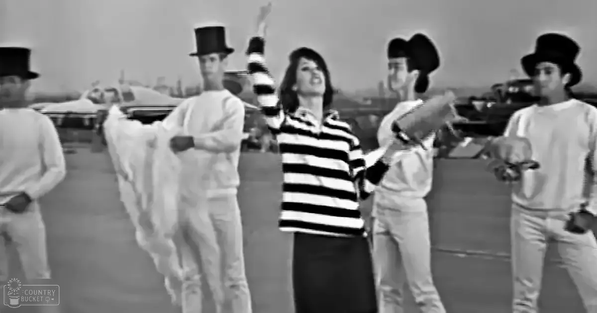 An adult female in a horizontally striped sweater stands amidst vintage cars on an open airfield with several middle-aged men holding rolled-up papers, suggesting a performance or event.