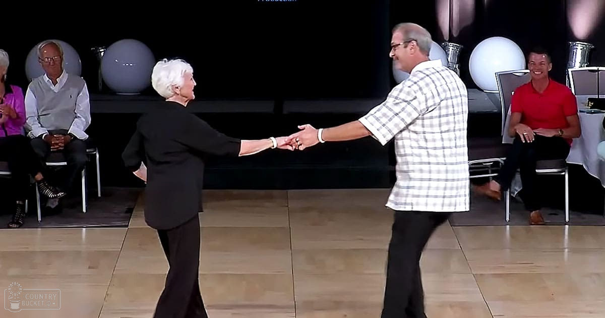 An older adult female in a black blazer and an adult male with glasses wearing a plaid shirt, seated among other dancers in what appears to be an indoor dance studio or event space.