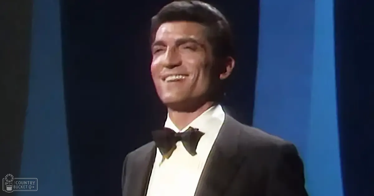 A young adult male in a tailored black tuxedo with white dress shirt and black bow tie, standing on stage with blue curtains behind him.