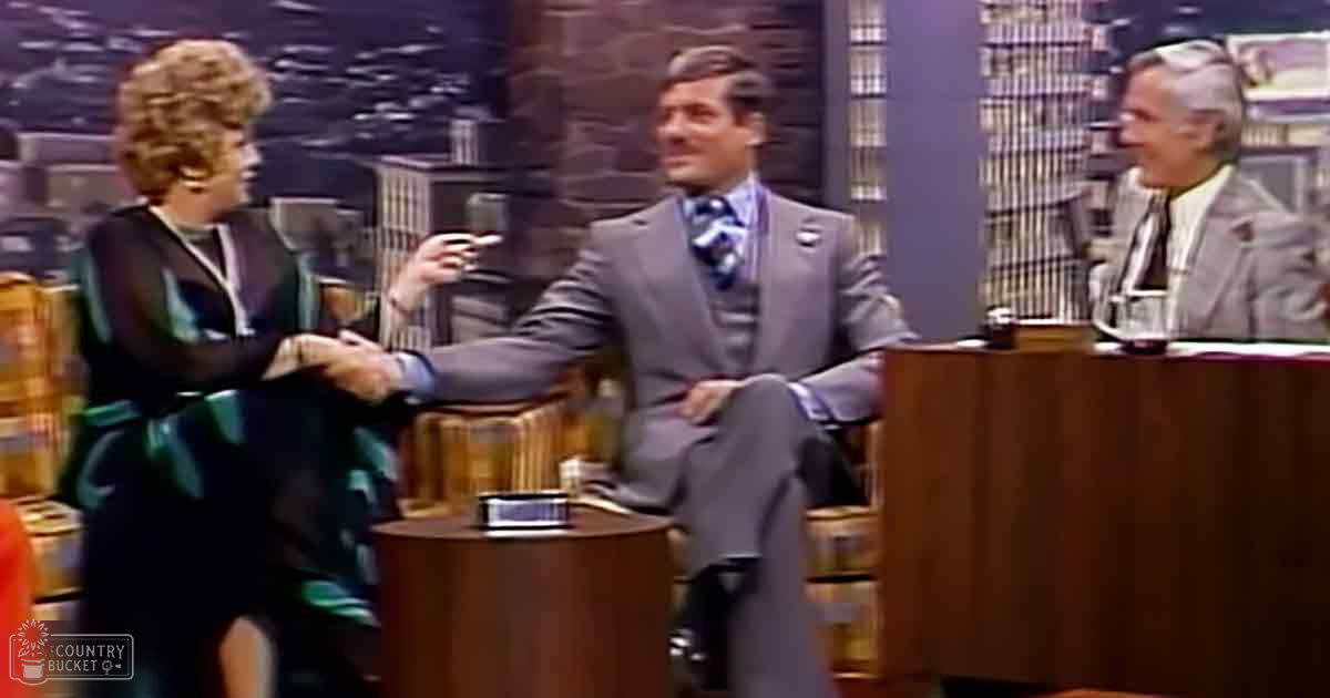 Talk show host and guests in a television studio with a cityscape backdrop.