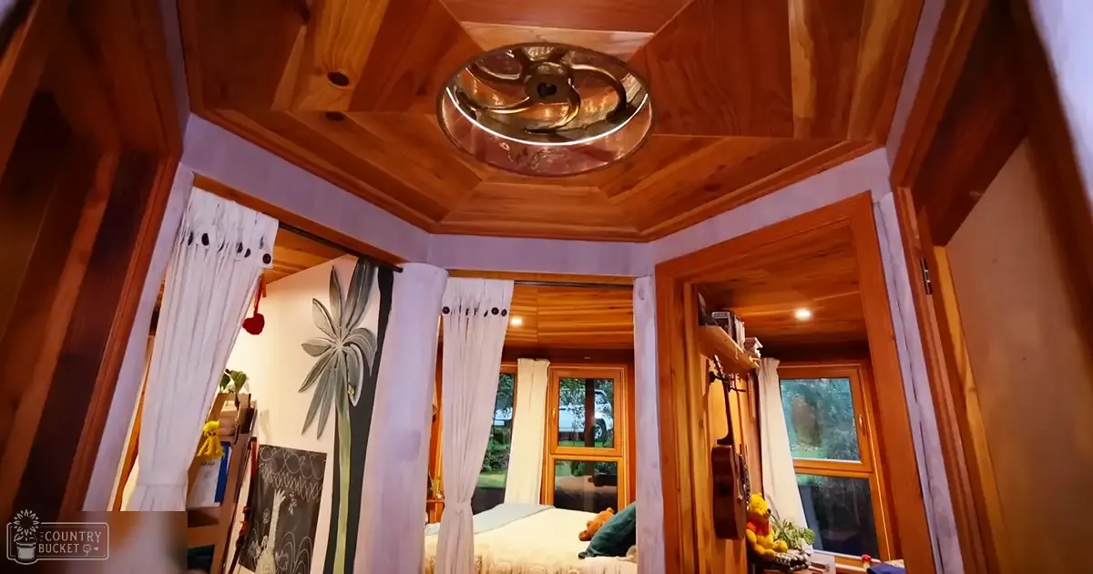 Cozy bedroom scene inside a tiny house, with a bed with floral-patterned curtains, a guitar on the wall, and a teddy bear near an open door leading outside. The setting is lush greenery visible through windows, suggesting spring or summer.