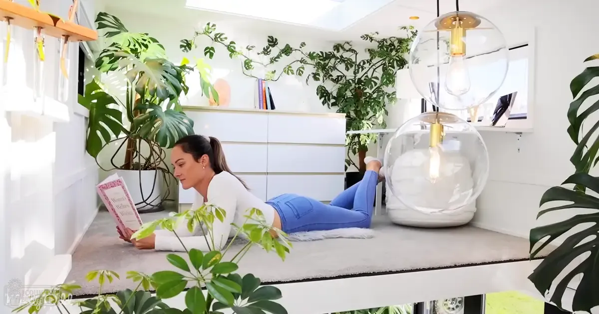 A young adult female reads on a white cushioned surface in an indoor setting with modern decor and lush potted plants.