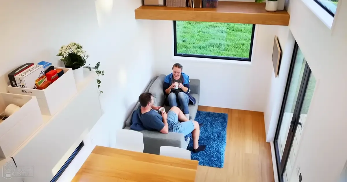A modern living space with a gray sofa, white chairs, a wooden dining table, and home decor including books and plants, viewed through large windows showing greenery. The room is brightly lit during spring or summer.