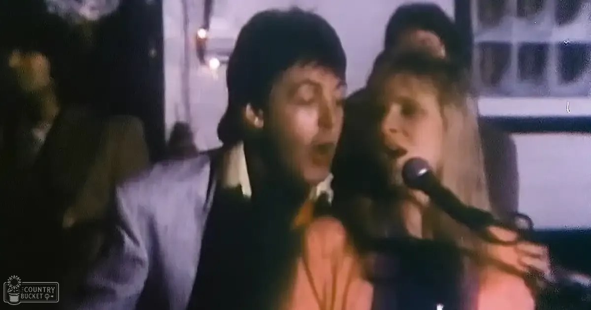 An adult male singer wearing a dark suit jacket and shirt, performing into a microphone with an adult female standing close behind in an indoor venue.