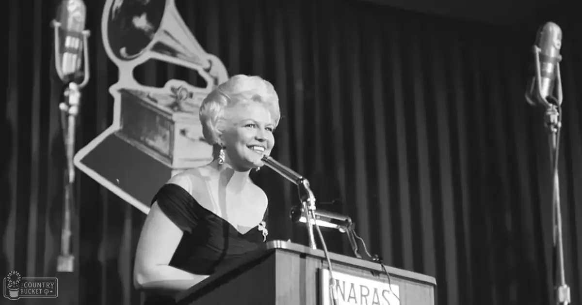 Middle-aged woman in a formal black dress at a Grammy awards ceremony with dark curtains and stage lighting behind her.