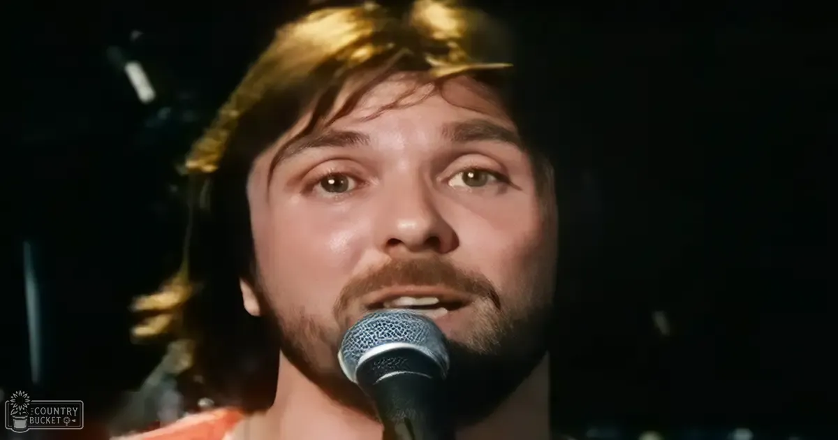A male adult wearing a casual, possibly vintage-style garment with an orange hue and textured pattern sings into a microphone in a dimly lit indoor setting designed for performances or recordings.