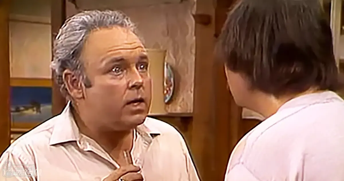 An older adult male wearing a light-colored collared shirt and tie, with another young adult male in similar attire, surrounded by domestic furnishings.