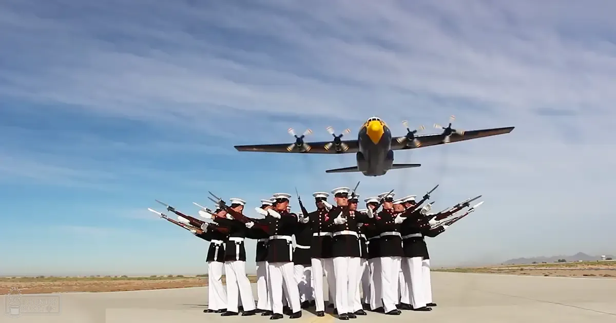 A group of young adult male soldiers in dark blue uniforms stand before a large military aircraft on an airfield under bright daylight, with distant hills or mountains visible.