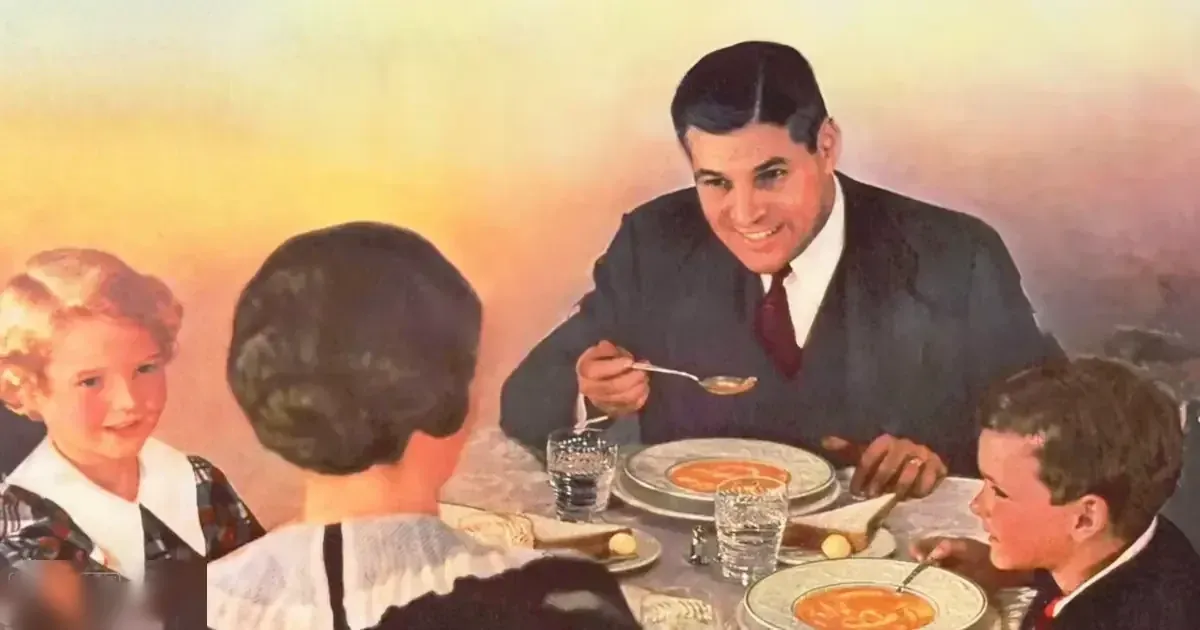 A family dining together at home in a warmly lit room, with adults and children wearing formal attire around a table set for soup and bread rolls.