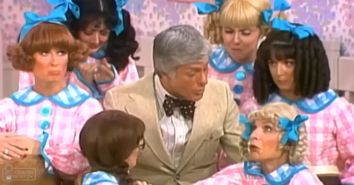 A middle-aged man in a light-colored suit with patterned shirt and bow tie, surrounded by young women in matching pink and white checkered outfits on a stage or set background.