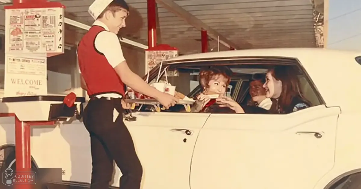 Teenage server in a red sleeveless shirt and black pants interacting with passengers at Sonic Drive-In, classic diner-style interior, warm weather, daytime natural light.