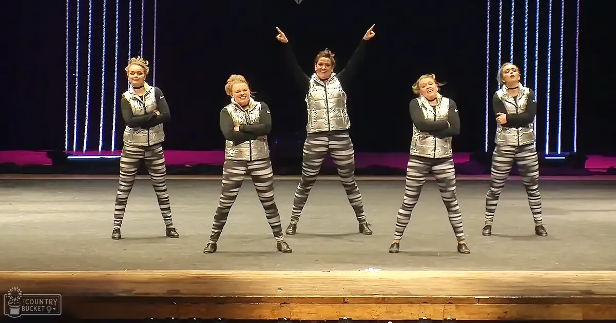 Young adult female dancers performing on an indoor stage with striped leggings, choker necklaces, and dark shoes under dramatic lighting.
