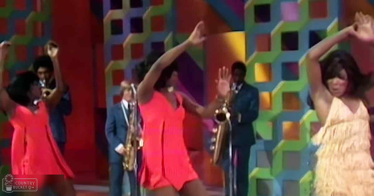 A vibrant performance scene with dancers in bright costumes and a colorful geometric backdrop, suggesting a lively 1960s-style setting.