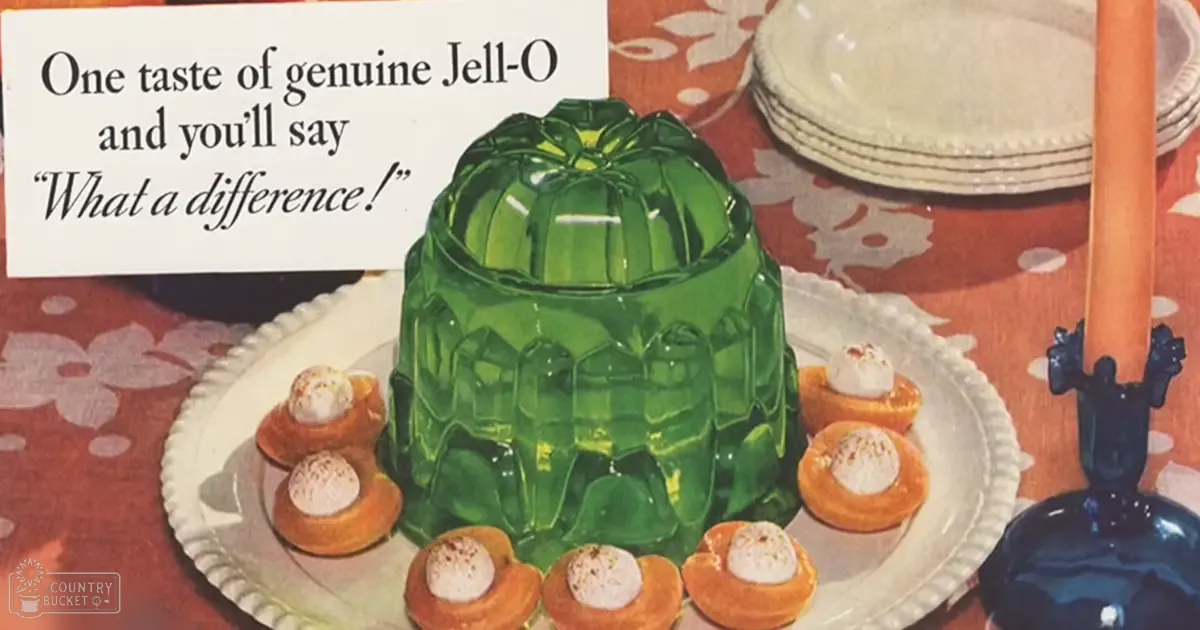 A large green Jell-O castle with small orange cream-filled cups arranged around it, placed on a white plate atop a red floral tablecloth in a dining room setting during the daytime.