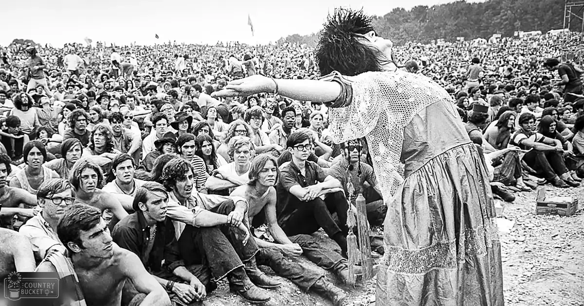 An adult male performer at Woodstock Festival with a patterned sleeveless top and textured skirt or dress surrounded by a crowd in casual summer attire, set outdoors during what appears to be late afternoon.