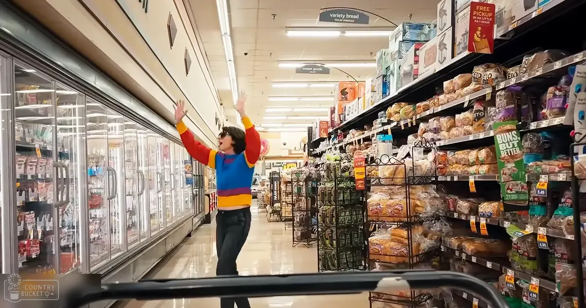 A young adult male wearing a long-sleeved, multicolored striped sweater with horizontal red, yellow, and blue stripes stands in an aisle of a grocery store filled with shelves stocked with baked goods. The setting is indoors under artificial lighting.