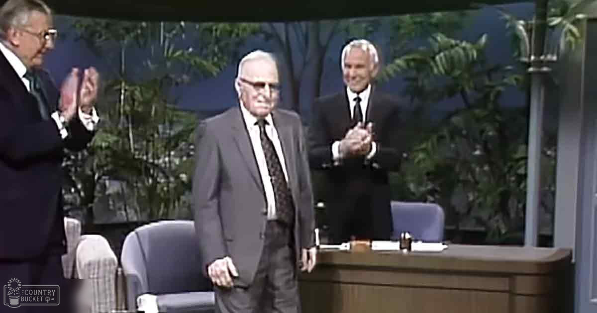 An older adult male singer in a dark suit with a patterned tie stands on stage during what appears to be a talk show set, surrounded by chairs and papers indicating a studio environment.