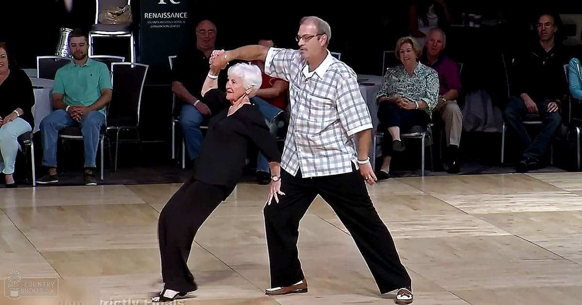 Adult male dancer wearing a plaid button-up shirt and black pants performs at an indoor dance event, surrounded by seated spectators under artificial lighting in a ballroom setting.