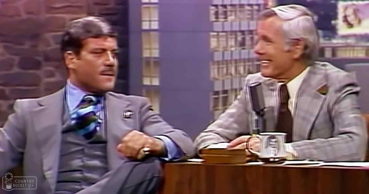 Adult male in a gray suit jacket and light blue dress shirt with patterned tie, seated on a talk show set with an older adult male wearing a light gray suit jacket over a white collared shirt; wooden desk, microphone, silver mug, and brick wall background.