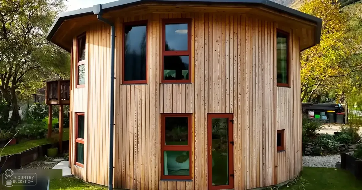 A small modern wooden cabin with vertical siding and red-framed windows, situated amidst a residential garden during autumn.