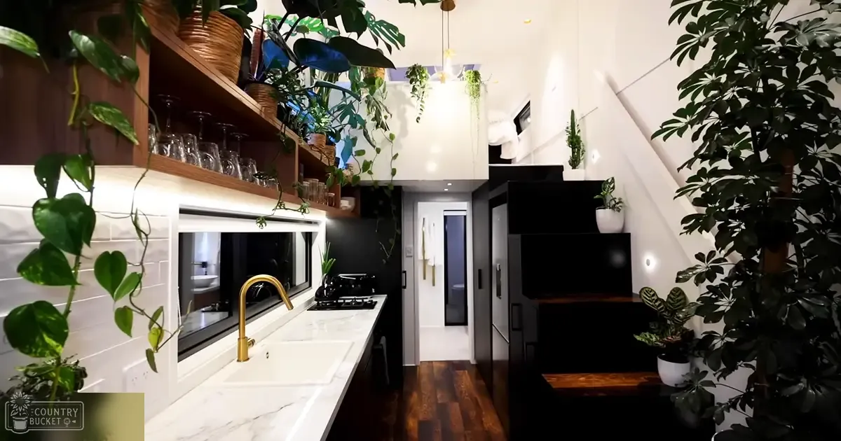 A modern kitchen with an open-plan layout in a cozy apartment or loft, with white marble countertops, a gold faucet, black stove with glass cooktop, built-in oven, wooden shelves holding glasses and plants, hanging light fixture, dark-colored refrigerator, slanted ceilings, natural daylight, white walls, black appliances, brown wood, green plants.