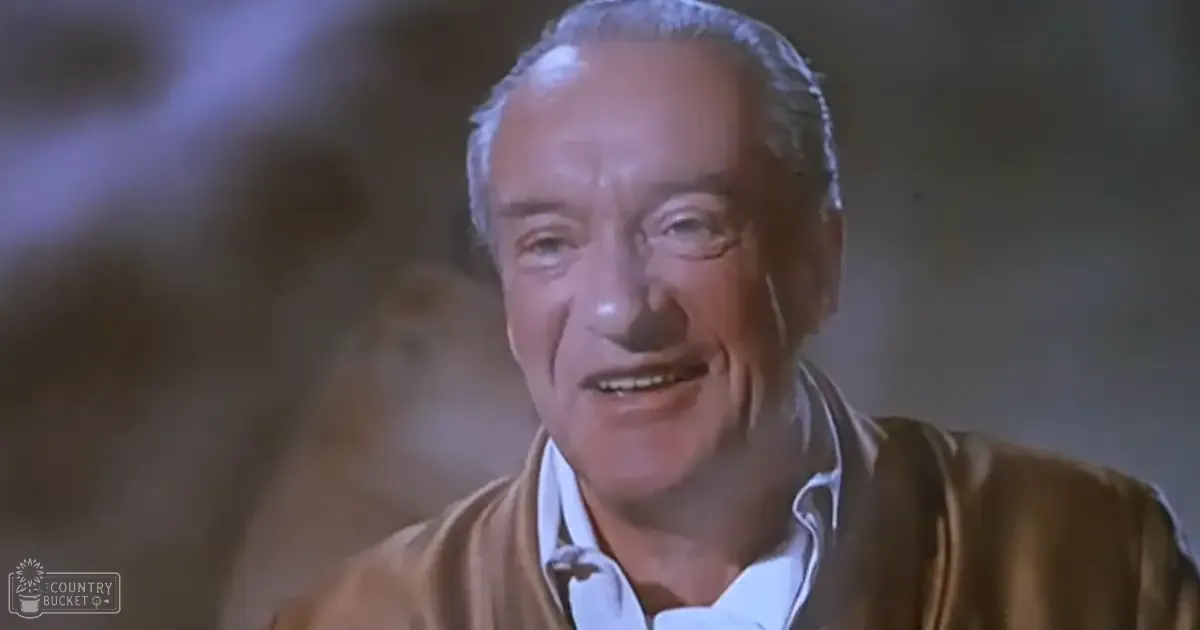 Older adult male in a film or television show scene, wearing an open-fronted brownish-gold garment with long sleeves under a collared white shirt with subtle pattern; setting appears to be indoors from an older film set with artificial lighting and muted background colors.